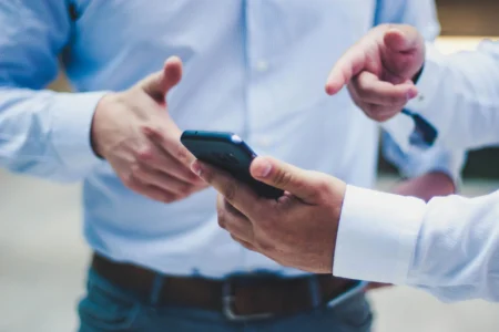 Zwei Männer in Geschäftskleidung im Gespräch, einer bedient ein Smartphone und zeigt mit seiner anderen Hand etwas auf dem Bildschirm, während der andere interessiert zuhört und seine Hand ausstreckt, als wolle er darauf reagieren.