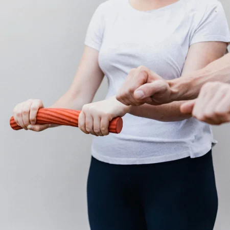 Zwei Personen führen eine Krankengymnastik-Übung mit einem roten Widerstandsband durch. Die eine Person hält das Band an beiden Enden, während die andere es in der Mitte hält und zieht, wobei die Anstrengung auf ihren Armen sichtbar ist.
