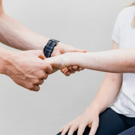 Eine Nahaufnahme zeigt eine physiotherapeutische Handgriff-Übung, bei der eine Person mit einem schwarzen Sportarmband den Unterarm einer anderen Person mit Sommersprossen hält. Der Hintergrund ist neutral und einfarbig.