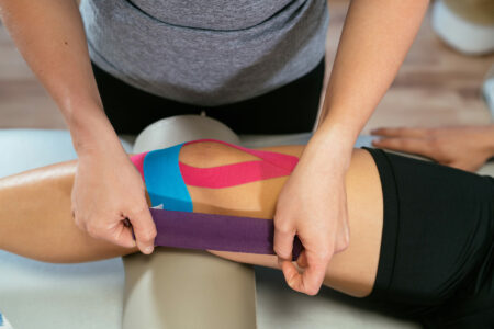 Eine Physiotherapeutin wendet farbenfrohes Kinesio-Tape am Knie einer Patientin an, um Schmerzen zu lindern und die Heilung zu unterstützen. Die Szene zeigt die Anwendung des Tapes in einem gut beleuchteten Behandlungsraum, was die moderne und professionelle Umgebung der Praxis widerspiegelt.