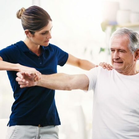 Eine Physiotherapeutin führt in einem hellen Raum eine Übung zur Schulterbeweglichkeit mit einem älteren männlichen Patienten durch. Sie unterstützt seinen Arm behutsam, während er lächelnd mitarbeitet. Die Praxis ist modern und gut ausgestattet, im Hintergrund sind Therapiegeräte sichtbar.